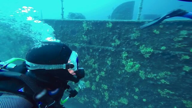 Diver Close Up Swimming Along Rusted Ship Wreckage - Diving Adventure in Slow Motion. Portrait of the diver with a mask, regulator, and exhaling bubbles, while swimming parallel to the historic wreck.
