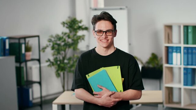 A young man with light skin and glasses stands in an office space. He holds vibrant folders and smiles. There are plants and office supplies around him, creating a productive environment.
