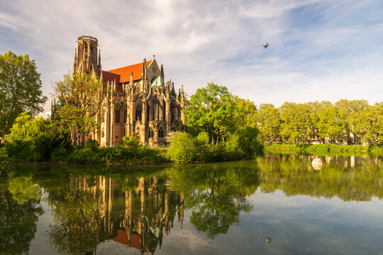 Spring sunlight on Johanneskirche by Feuersee lake with clear reflections, green trees, and calm water in Stuttgart, Germany.
