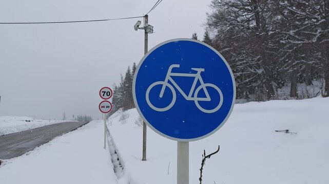 Blue circular road sign indicating a bicycle route stands beside an empty road. The surrounding landscape is covered in fresh snow during a winter snowfall