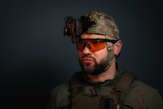 Bearded military man wearing a tactical night vision cap with a monocular NVG device attached in a flipped up position against a dark background