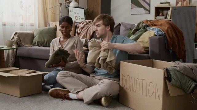 Diverse gen Z couple packing clothes into cardboard boxes for donation while sitting on floor after decluttering their wardrobes, with couch in background