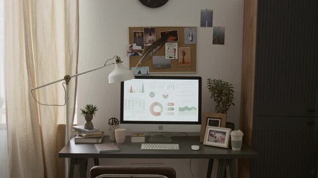 No people view of wooden desk featuring computer monitor displaying colourful charts, keyboard and mouse, books, small potted plants, lamp and photo frames, also cork board on wall