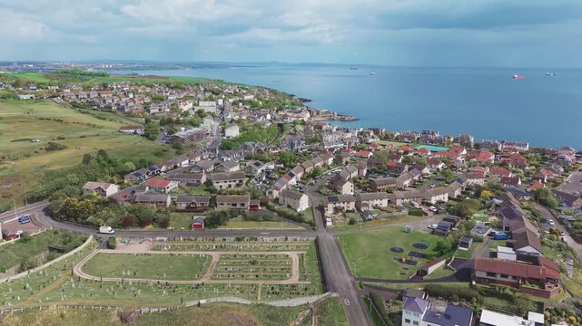 Aerial cinematic drone forward flyover shot starting over historic cemetery and residential streets in Pettycur, moving towards the scenic sea coast with cargo ships on the horizon, Fife, Scotland.
