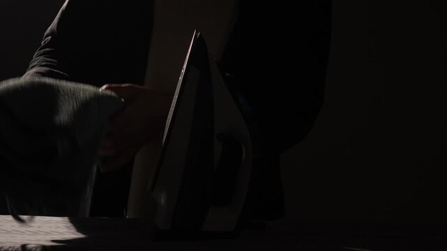 Man's silhouette visible in low light, hands holding clothes and operating an iron on an ironing board, performing routine household laundry tasks in a dark room
