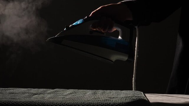 Person's hand gripping a modern steam iron, releasing vapor as it glides over a textured dark fabric on an ironing board, highlighting daily household chores and domestic life
