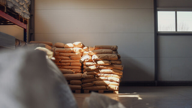 Stacks of large paper sacks on wooden pallets in a bright industrial warehouse interior with natural sunlight from a window