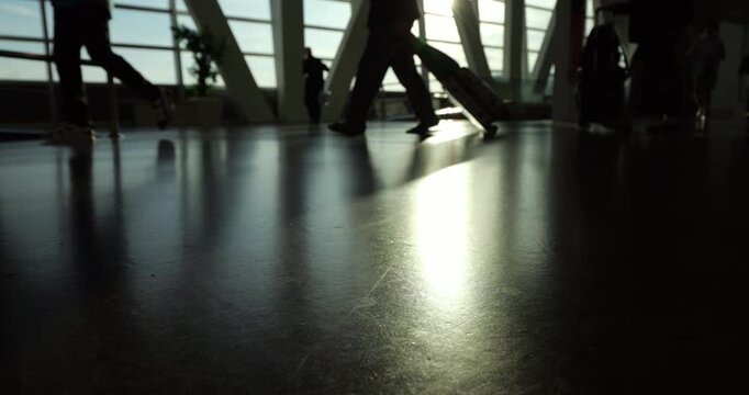 High-contrast shot of spacious passage at airport, focus on floor at foreground with blurred silhouetted passenger legs and rolling luggage move through corridor.
