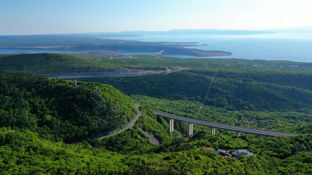Panoramic aerial view of the lush Croatian coastline, featuring the iconic Krk Bridge spanning the Adriatic Sea, with winding roads and vibrant green hills. Highway A7