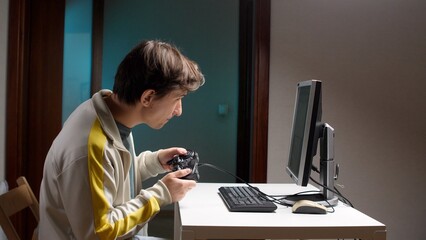 Young man intensely focused on gaming, seated at a desk with a wired controller, immersed in an online video game on a computer monitor in a modern home setting © Aliaksandr