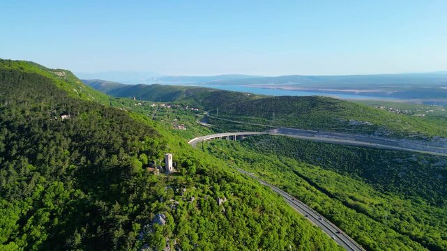 Aerial view of Croatia's lush green hills, winding highway A7, and the stunning Kvarner Gulf with islands under a clear spring sky.