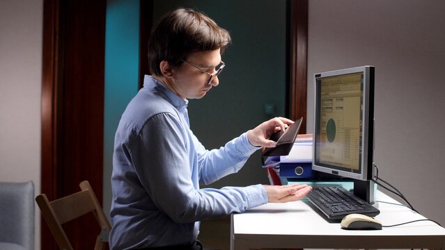Man at home office desk stares into an open empty wallet while computer displays declining financial data, conveying stress, debt worries and budget crisis during recession
