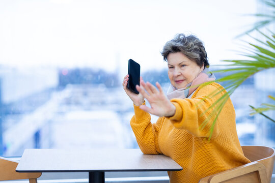 Mature woman in yellow sweater speaking on mobile phone, gesturing with hand, emotional conversation in modern interior.