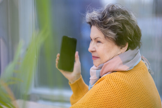 Mature woman in yellow sweater speaking on mobile phone, gesturing with hand, emotional conversation in modern interior.