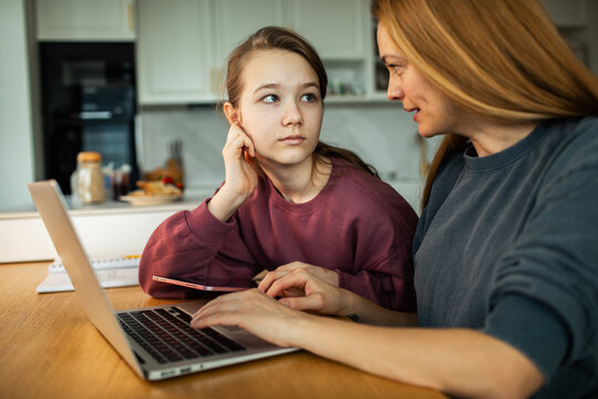 Mother helping teenage daughter with homework on laptop at kitchen table