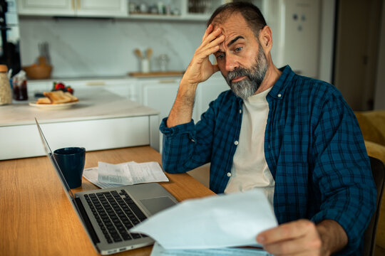 Stressed middle-aged man reviewing bills at home kitchen