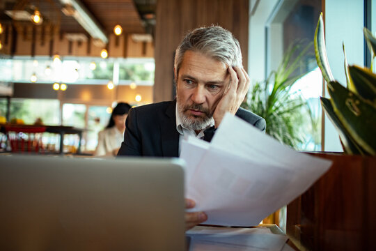 Stressed senior businessman reviewing bills on laptop in cafe