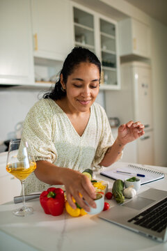 Smiling woman cooking vegetables with laptop in home kitchen