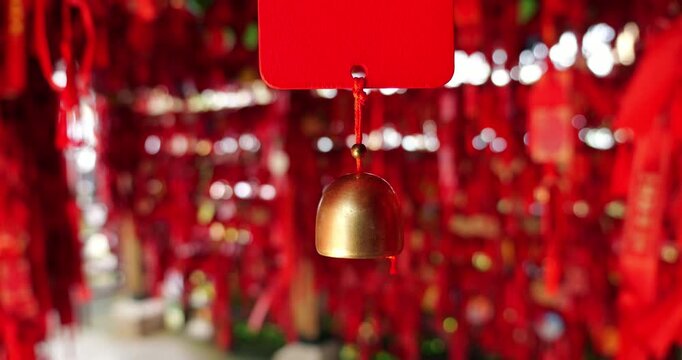 Close-up shot focused on single golden brass bell hanging from red prayer tag, swaying gently in slow motion. Blurred background features thousands of vibrant red ribbons and wishing tags