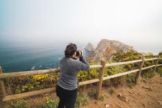 Woman standing on a cliff viewpoint capturing the dramatic rock formations and the vast blue sea with her camera, enjoying travel and the scenic overlook in Asturias, Spain