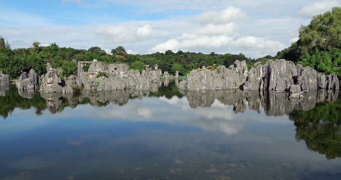 Panning shot shows scenic limestone formations of Shilin perfectly reflecting in calm waters of Shilin Lake. Towering grey karst pillars contrast against lush green trees and soft white clouds