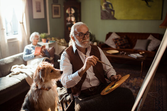 Senior man painting on easel at home, dog watching
