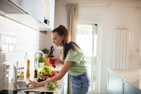 Woman preparing salad and using laptop in home kitchen
