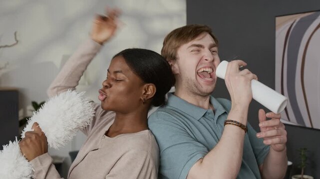 Chest-up shot of positive diverse gen Z couple pretending to sing into feather duster and spray bottle while doing housework