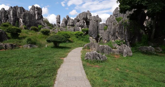 First-person view walking along paved stone path toward towering karst rock formations at Shilin Stone Forest. Moving from shuttle bus terminal stop, dense area of jagged limestone pillars ahead