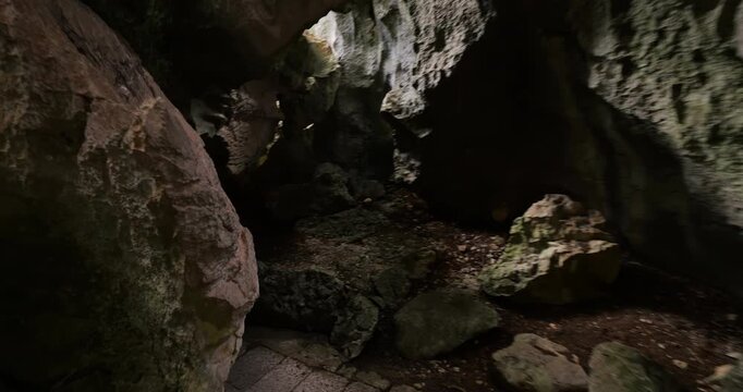 First person perspective, walking through narrow, shadowed path between rough limestone rocks and uneven karst walls. After short corridor, camera turns left and emerges toward sunlit exit