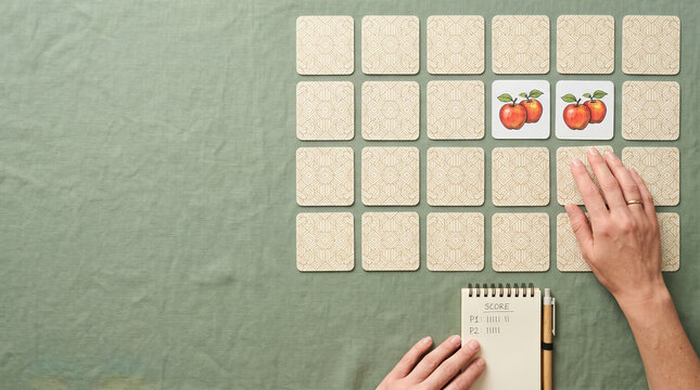Woman playing memory game with matching apple cards on green tablecloth. Tabletop concentration board game with flat lay layout and score pad for recreational family home fun activity.