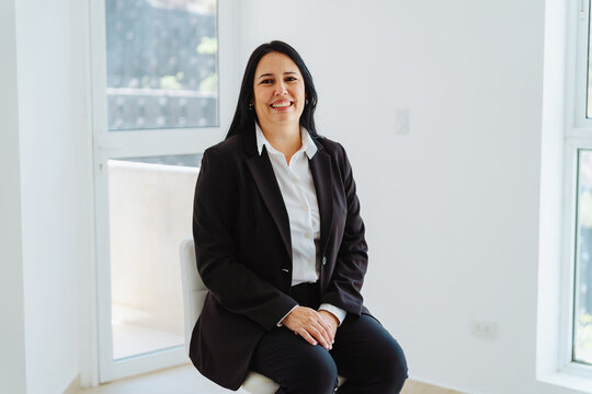 Confident Latin woman real estate agent sitting in an empty room, presenting a new property with a welcoming smile