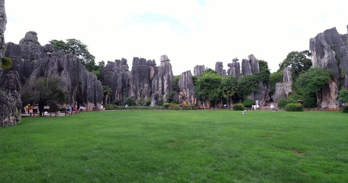 Panoramic pan across green meadow circled by majestic limestone karst formations at Shilin Stone Forest, Yunnan, China. Rugged grey stone pillars, some capped with precarious balancing boulders