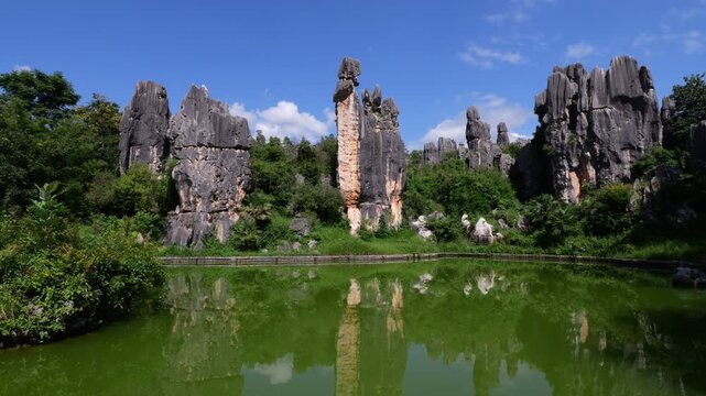Cinematic shot moves forward along stone-paved embankment, tilting up from green waters of tranquil pond. Smooth motion reveals legendary Ashima Rock standing tall as figurative centerpiece