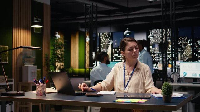 Businesswoman doing accounting task and taking notes at office desk, using laptop to transcribe and evaluate operational metrics. Staff member working late to meet objectives for strategy.