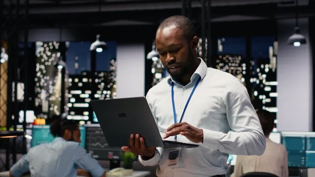 African american man, walking with laptop, office professional evaluating operations. Finishing tasks with notebook, Business manager employee working late at night to resolve complaints.