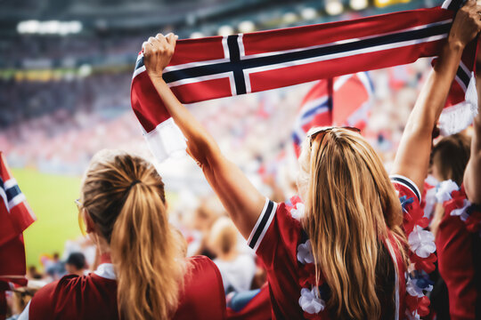 Back view of two female supporters holding the Norway national flag overhead during a soccer match in a crowded stadium atmosphere.