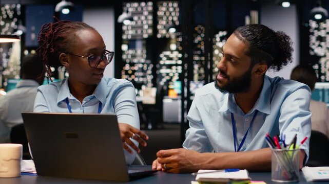Diverse team of businesspeople at desk using laptop evaluating metrics, teamworking concept late at night. Colleagues working late at night on notebook to assist online clients, finishing tasks.