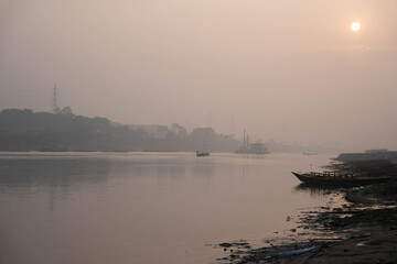 Naklejka premium A serene landscape of a riverbank at sunset with a small wooden boat moored on the muddy shore under a hazy, golden sky in rural bangladesh