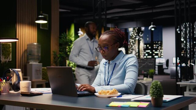 Eating disorder and emotional stress, black woman working on laptop, worker eating excessive junk food at her desk. Employee appearing worried as she handles tasks, compulsive overeating.