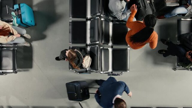 Travelers in airport lounge sitting on chairs, holding travel documents. Passengers with luggage and boarding passes waiting for aircraft in air transportation hub, top down view shot