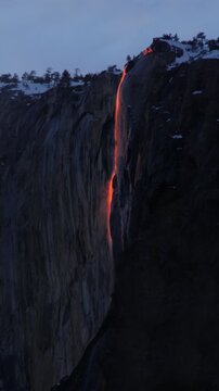 Vertical Screen: Yosemite National Park Firefall phenomenon with glowing orange waterfall cascading down massive granite cliff face at dusk with snow-capped peaks and pine trees silhouetted against tw