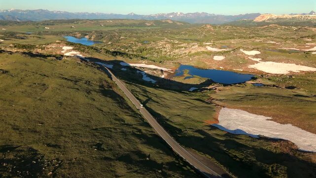 Drone footage follows a white vehicle traveling along Beartooth Highway in Montana through stunning alpine terrain with blue lakes and melting snow patches amid green meadows.
