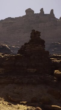 A large rock formation stands in a dry area surrounded by hills. The sun shines down on the scene creating strong shadows on the ground. No plants or water are visible in the landscape.