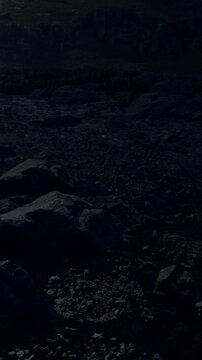 A rocky landscape is displayed at sunset with dark stones scattered on the ground. The mountains rise in the background under a dim sky. The scene captures nature&rsquo;s raw beauty.