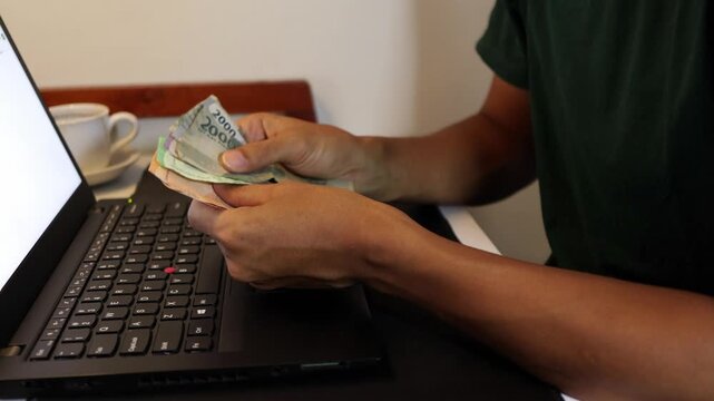 Close up of a man's hands counting Indonesian Rupiah banknotes in front of an open laptop at a desk.