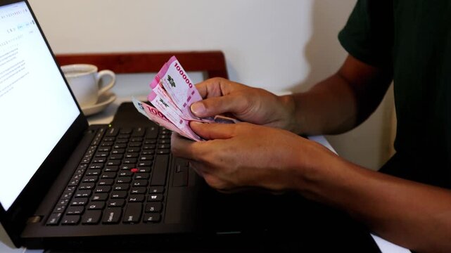 Close up of a man's hands counting Indonesian Rupiah banknotes in front of an open laptop at a desk.