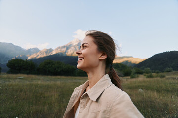 Fototapeta premium Woman lifestyle portrait outdoors in open fields with mountains, smiling and relaxed, wearing light jacket, enjoying natural scenery and warm sunlight.