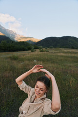 Fototapeta premium A happy woman in casual beige clothing enjoys nature with raised arms in a green meadow near forested hills under a clear sky. Lifestyle and outdoor concept.