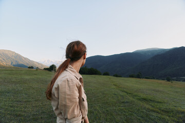 Fototapeta premium Woman seen from behind in a casual jacket stands in a lush field with distant mountains, creating a serene outdoors lifestyle scene focused on nature and exploration